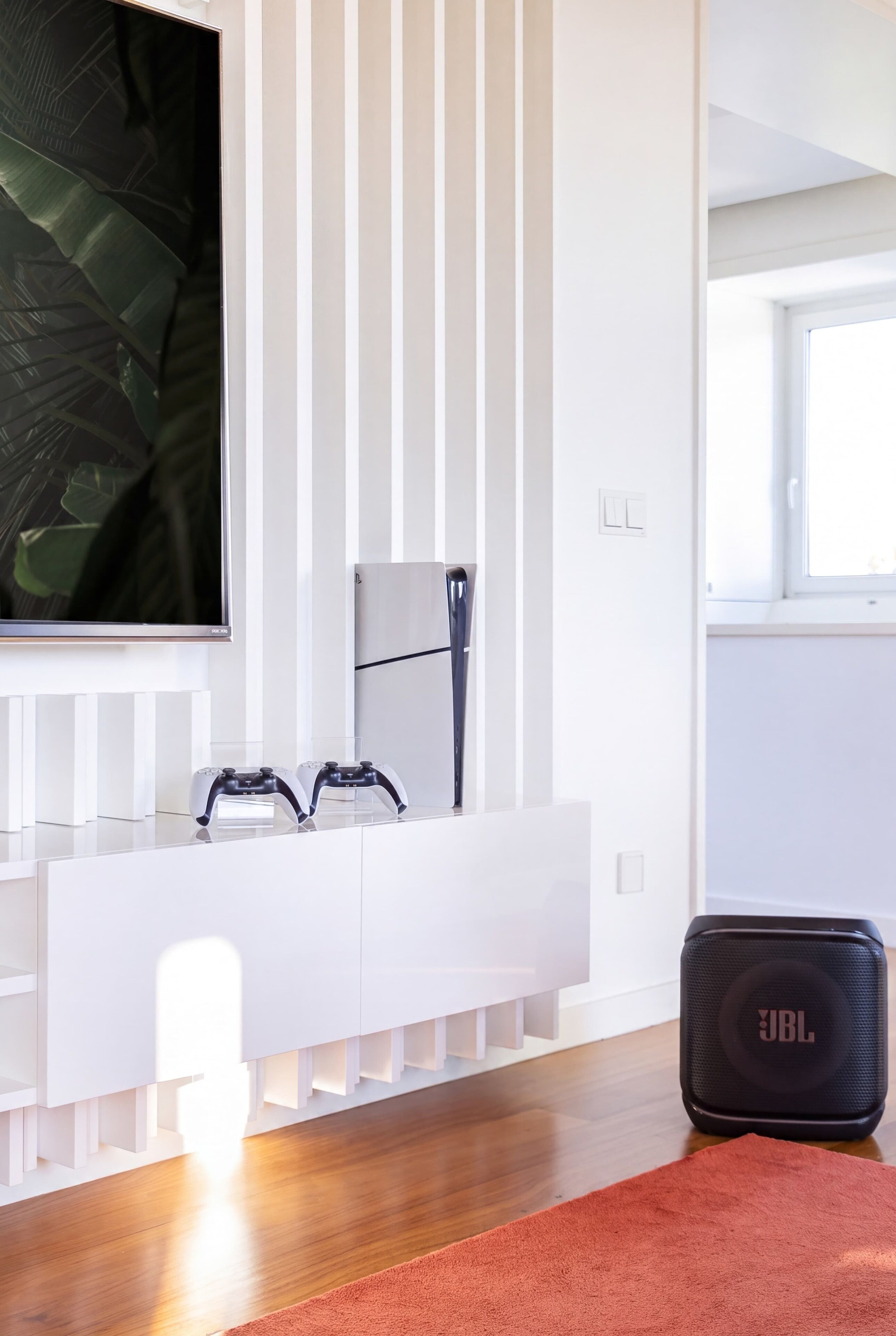Ein modernes Zimmer mit einem weißen Schminktisch, zwei Spielcontrollern, einem großen JBL-Lautsprecher und einem großen Fenster.