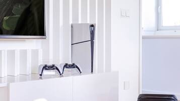 Ein modernes Zimmer mit einem weißen Schminktisch, zwei Spielcontrollern, einem großen JBL-Lautsprecher und einem großen Fenster.