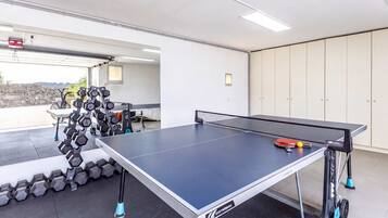 Ein Tischtennis-Tisch mit Netz, eine Hantelbank und ein Fahrrad in einem Raum mit Fliesenboden und einer zur Außenseite führenden Tür.
