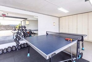 Una mesa de ping pong con red, un juego de mancuernas y una bicicleta en una habitación con piso de baldosas y una puerta que da al exterior.