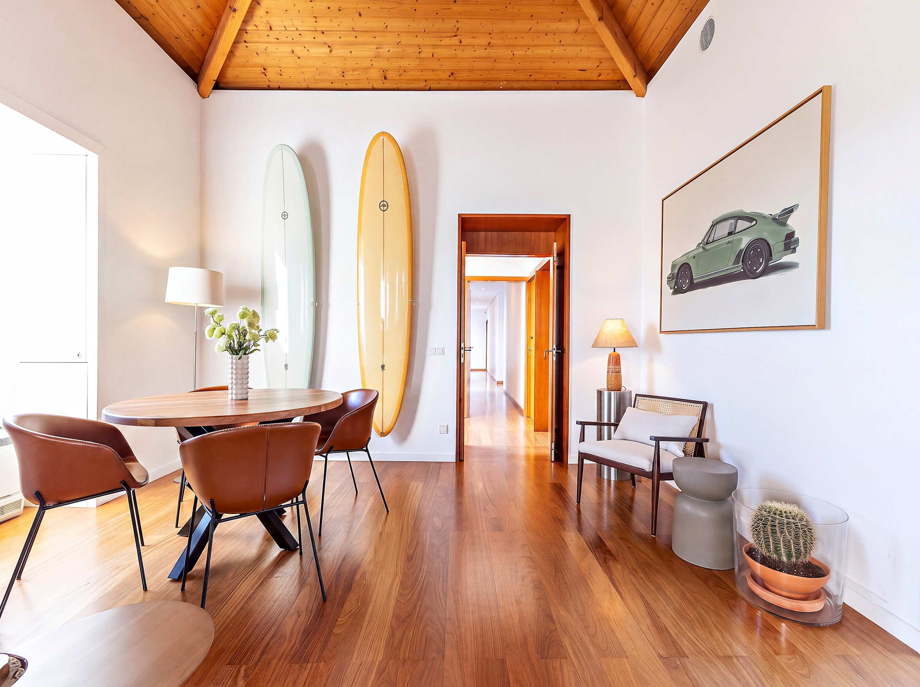 Ein Zimmer mit Holzboden, einem Esstisch mit Stühlen, einer Lampe, einem gerahmten Bild an der Wand und zwei Surfbrettern, die an die Wand gelehnt sind.