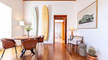 Ein Zimmer mit Holzboden, einem Esstisch mit Stühlen, einer Lampe, einem gerahmten Bild an der Wand und zwei Surfbrettern, die an die Wand gelehnt sind.