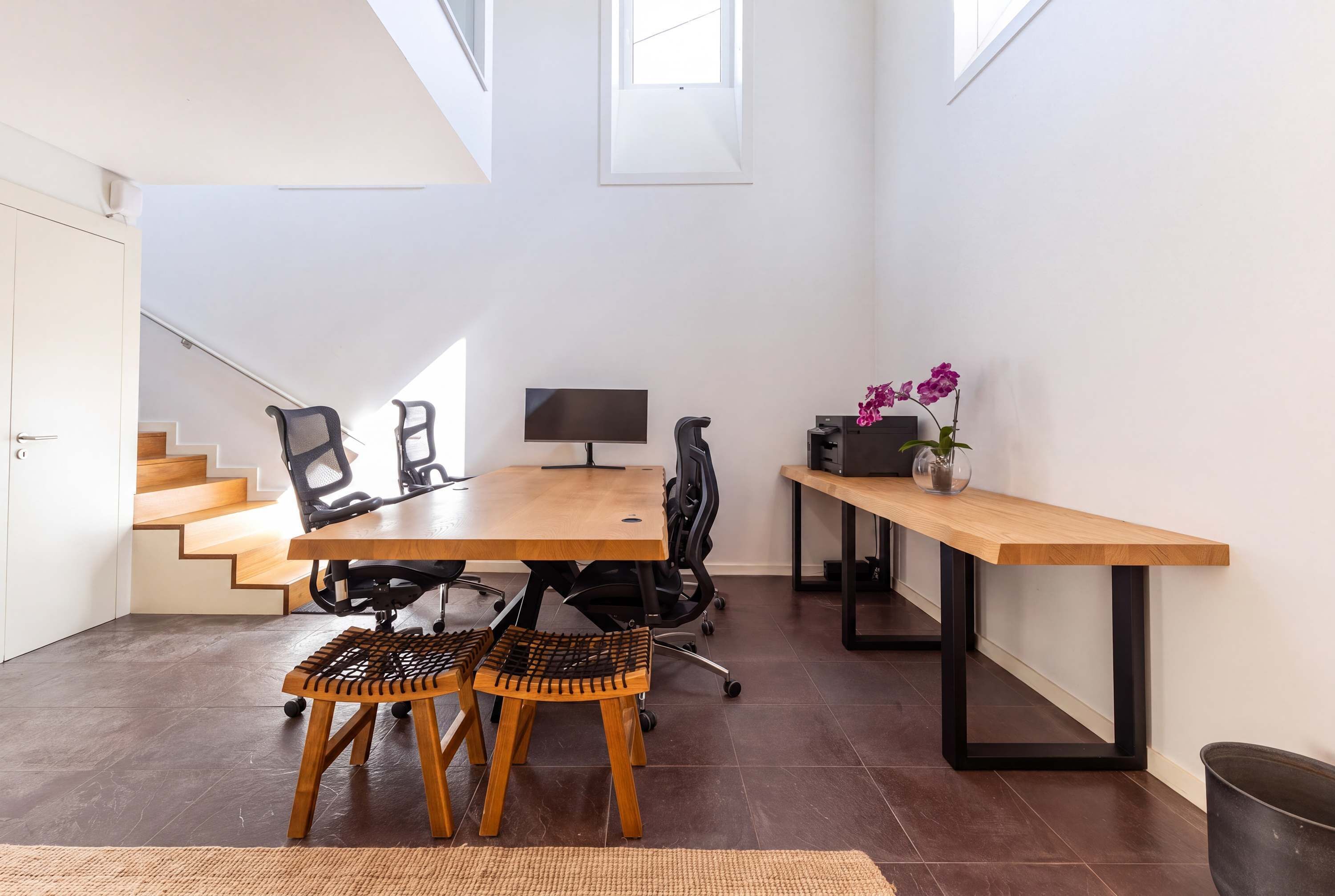 A modern office space with a wooden table, office chairs, a computer, and a small desk with a vase of flowers.