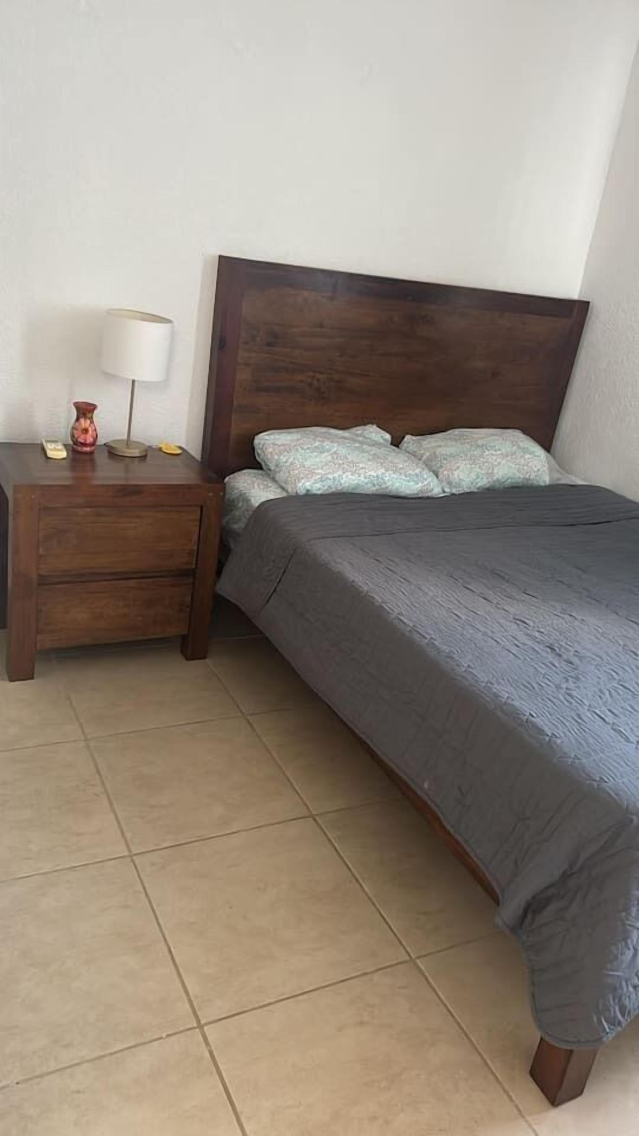 A bedroom with a wooden headboard, a bed with grey bedding, a bedside table with a lamp, and a small decorative item.