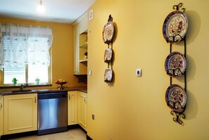 A kitchen with yellow walls, a dishwasher, a window with sheer curtains, and a fruit bowl on the counter.