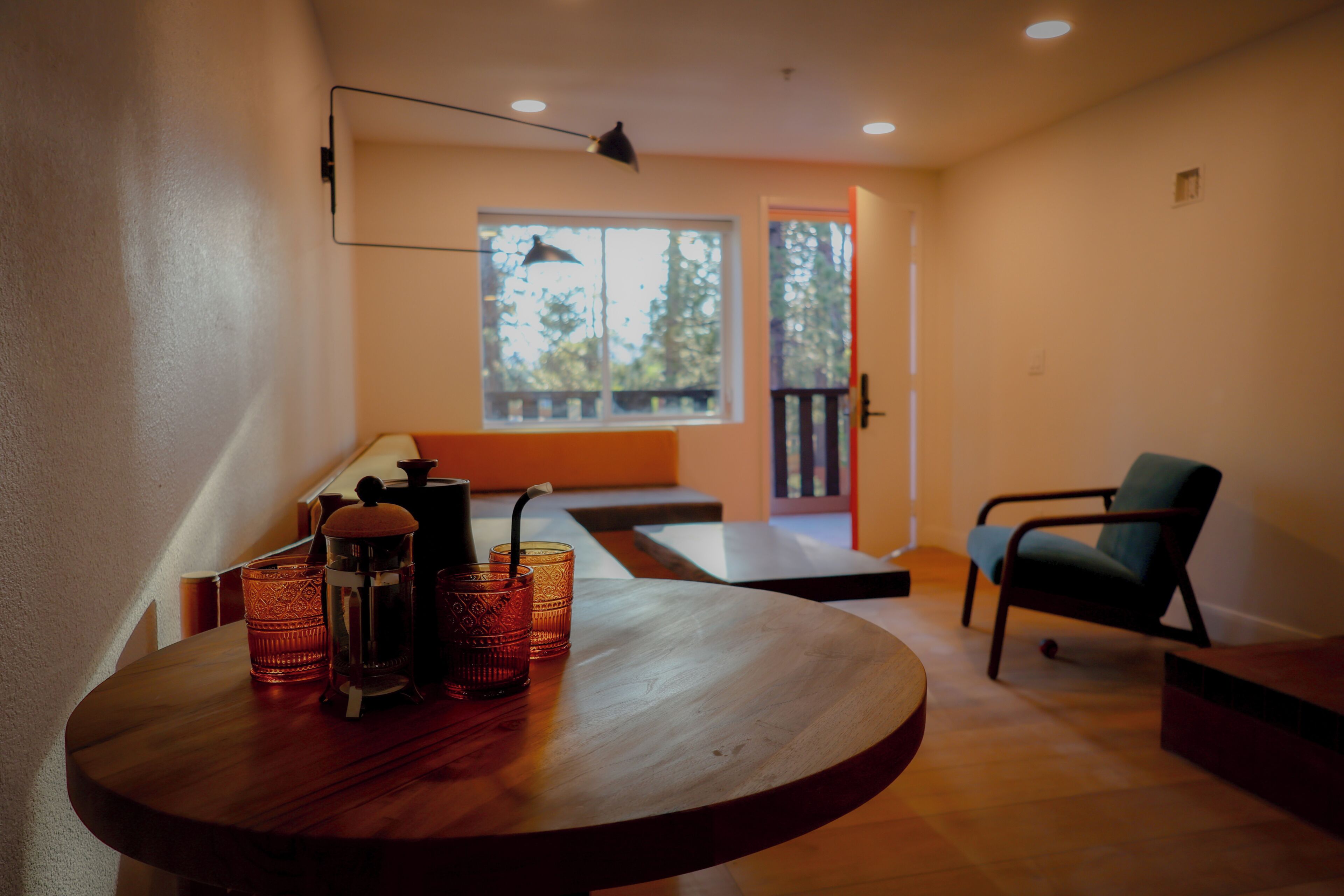 A room with a round wooden table, a chair, a window with a view of trees, and a door leading to an outdoor area.