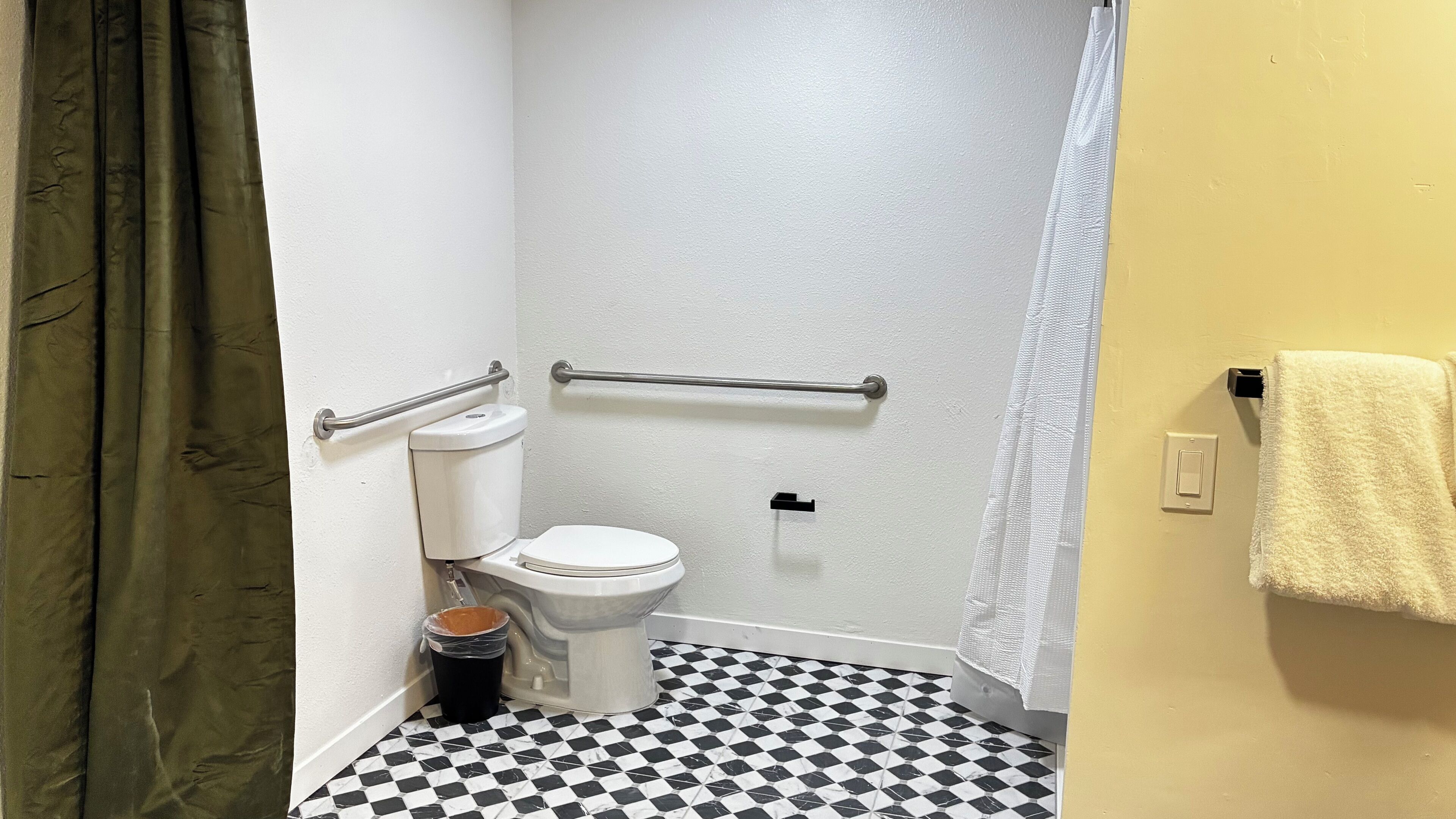 Une petite salle de bain comprenant des toilettes, une barre d’appui, un rideau de douche, un porte-serviettes et une poubelle.