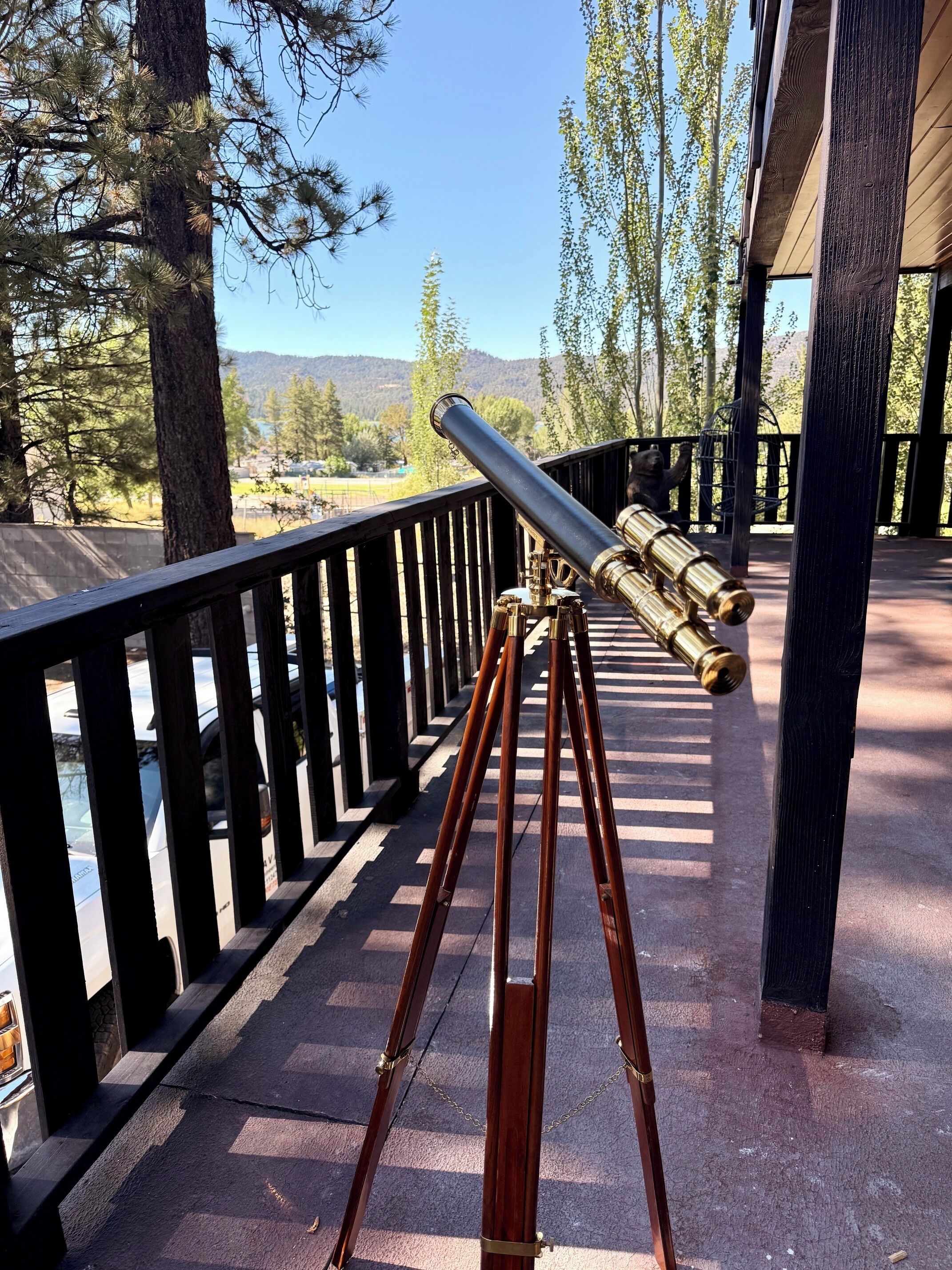Un télescope, posé sur un trépied en bois, se trouve sur un balcon doté d’une balustrade, d’où l’on peut admirer un paysage arboré et une vue lointaine.