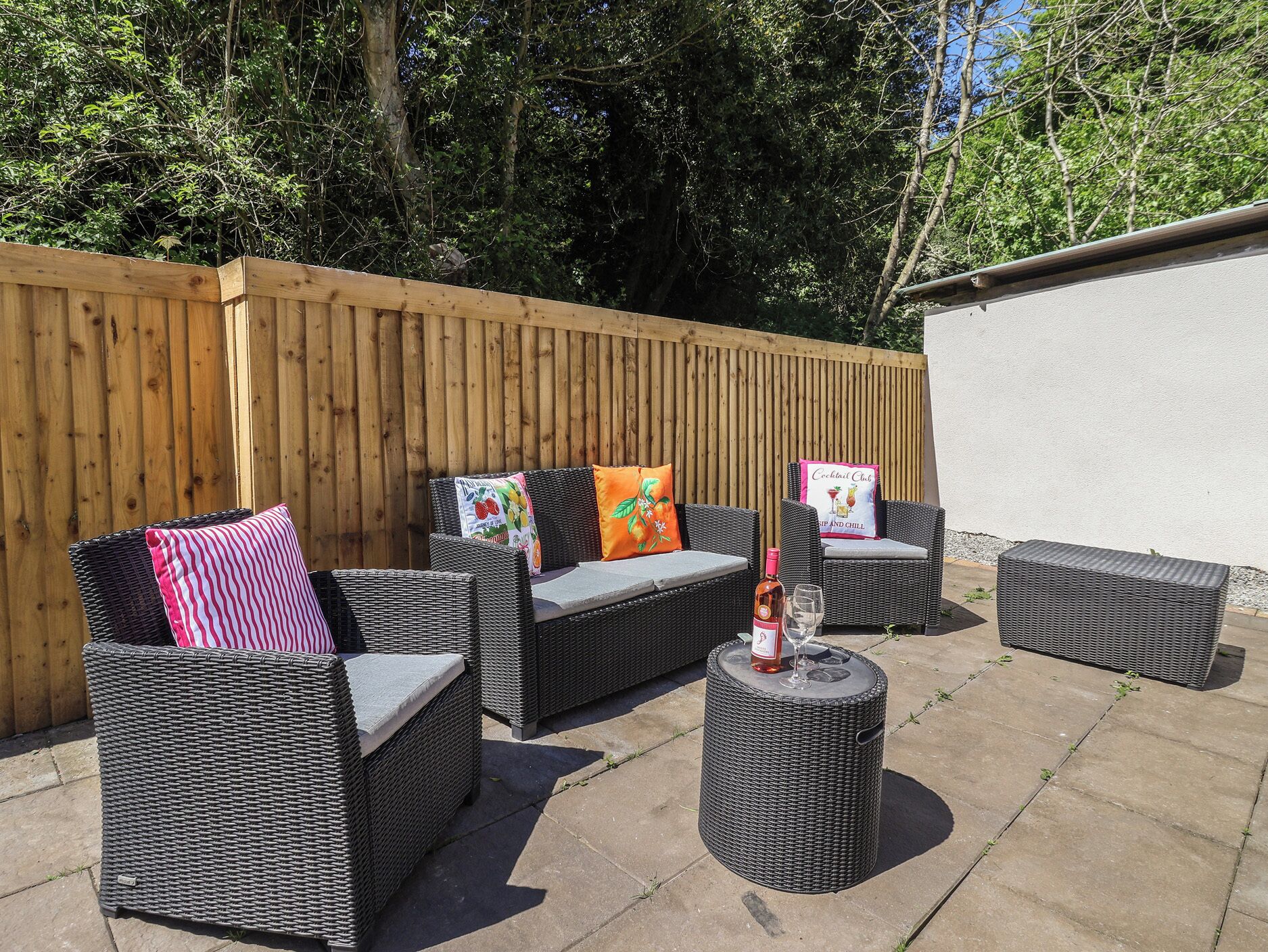 An outdoor seating area with wicker furniture and a small round table, surrounded by a wooden fence and a white wall.