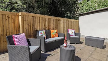 An outdoor seating area with wicker furniture and a small round table, surrounded by a wooden fence and a white wall.