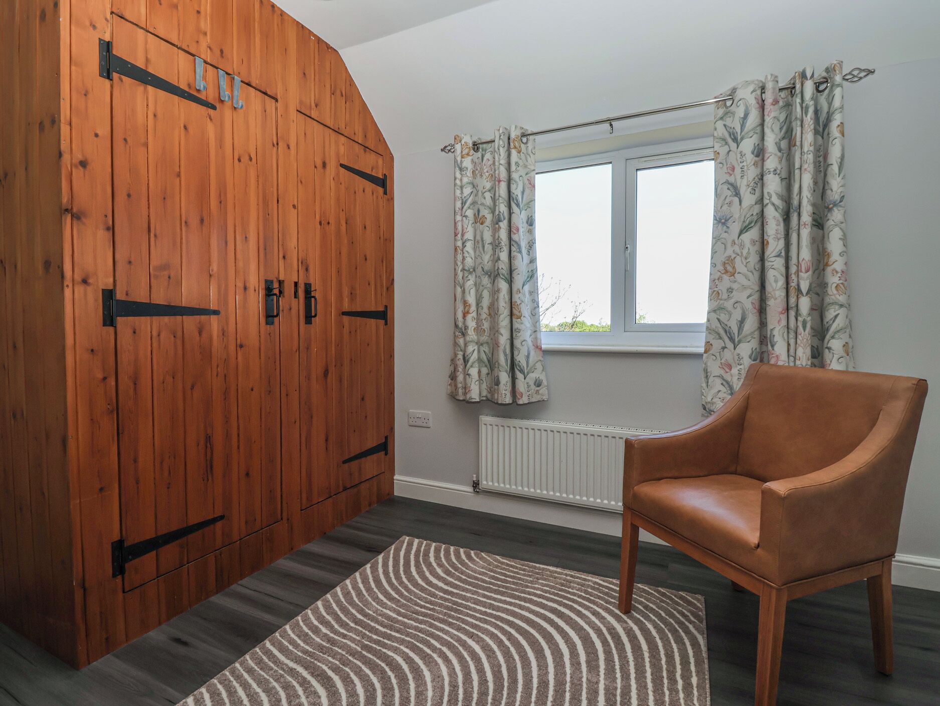 A room with a wooden wardrobe, a window with floral curtains, a radiator, and a leather chair.