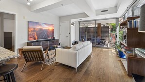 A modern living room with a white sofa, wooden flooring, a large painting, and a dining area with a granite table. - 4 MONTH MINIMUM RENTAL-Cozy 1 bedroom and 1.5 bath in the Iconic Optima ! (Scottsdale)