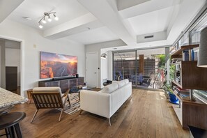 A modern living room with a white sofa, wooden flooring, a large painting, and a dining area with a granite table. - 4 MONTH MINIMUM RENTAL-Cozy 1 bedroom and 1.5 bath in the Iconic Optima ! (Scottsdale)