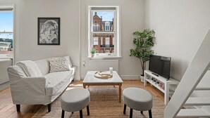 Una sala de estar moderna con sofá, mesa de centro, reposapiés, mueble para televisor y ventana con vista a un edificio.