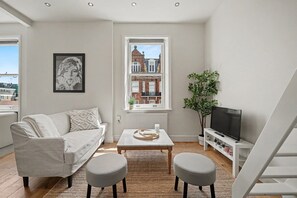 Ein modernes Wohnzimmer mit Sofa, Couchtisch, Hocker, TV-Schrank und einem Fenster mit Blick auf ein Gebäude.