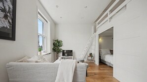 Una habitación pequeña con un sofá, un televisor, una planta y una escalera que sube a un dormitorio.