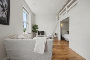 Ein schmales Zimmer mit einer Couch, einem Fernseher, einer Pflanze und einer Treppe, die in ein Schlafzimmer führt.
