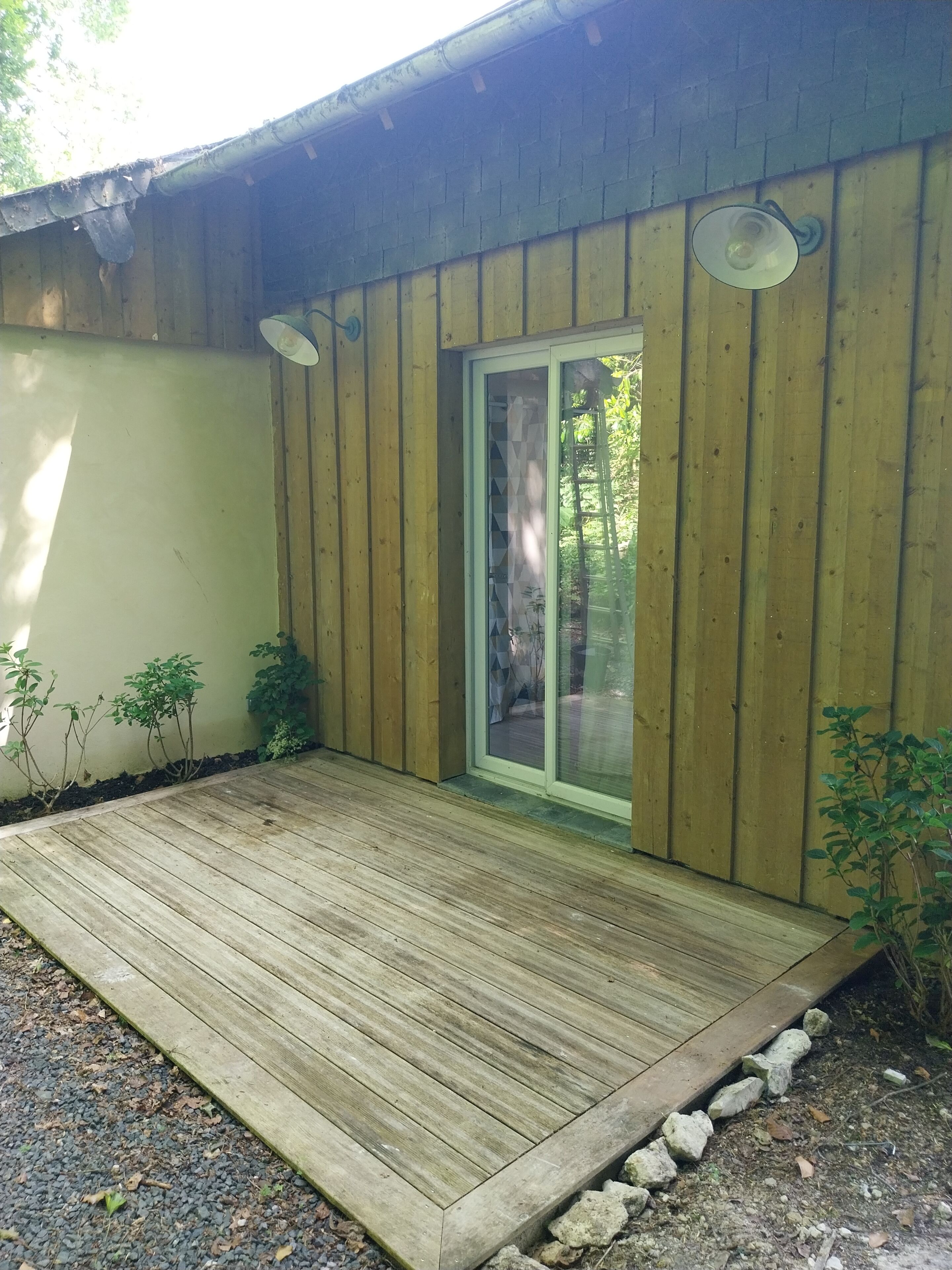 A wooden deck with a glass door leading to a covered area, surrounded by small plants and a gravel path.