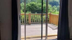 Une pièce dotée d’une porte-fenêtre coulissante donnant sur une terrasse extérieure avec une rambarde en bois, offrant une vue sur une colline boisée et dotée d’un sol carrelé.