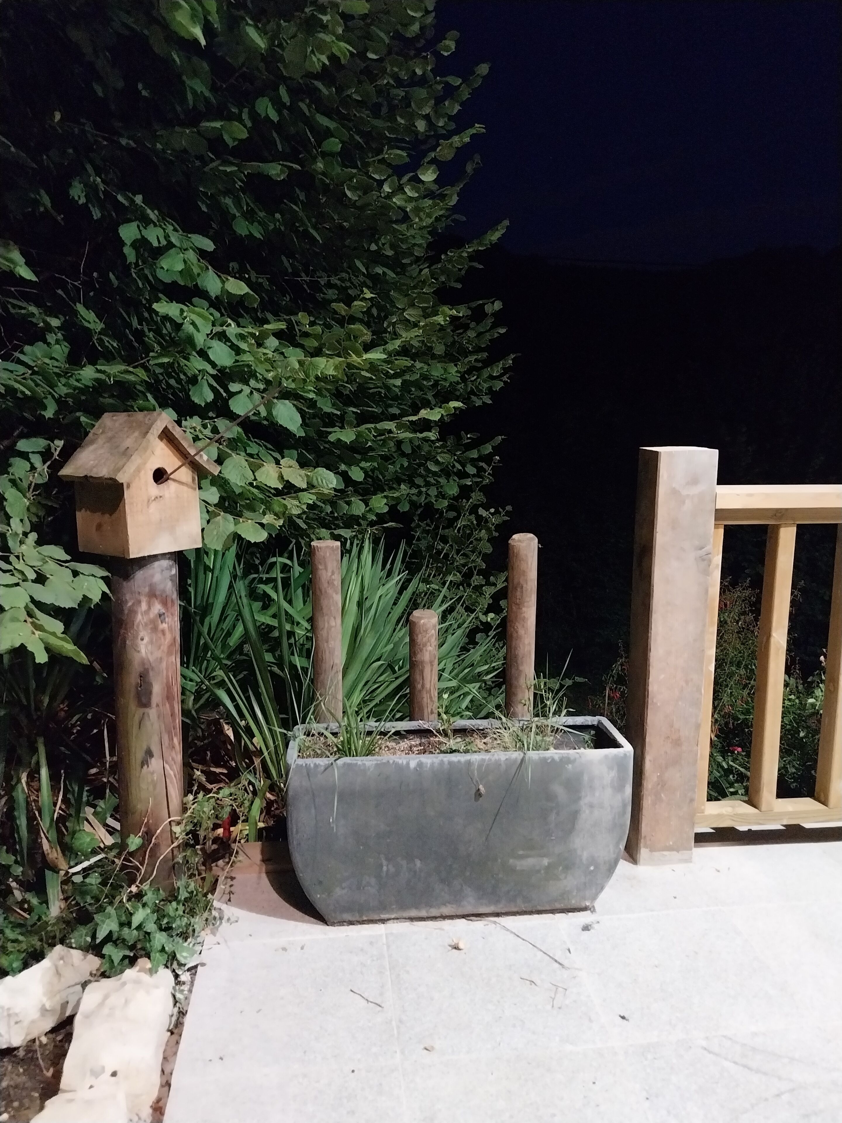 Uma casa de pássaro de madeira presa a um poste, um cachepot de concreto com plantas e uma cerca de madeira.