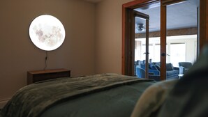 Un dormitorio con una cama, una lámpara de pared redonda y vistas a un salón a través de una puerta de cristal.