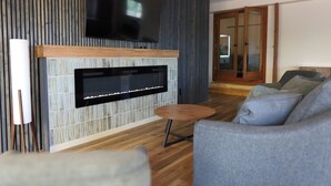 Una sala de estar moderna con un televisor de pantalla plana montado en una pared con textura, una chimenea, una mesa redonda de madera y un sofá gris.