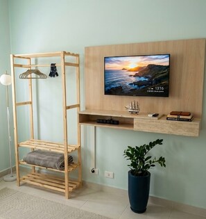A room with a TV mounted on a wooden wall, a wooden shelf below it, a hanging rack, a potted plant, and a floor lamp. - Refúgio exclusivo frente mar no Gonzaga em Santos/SP (Santos)