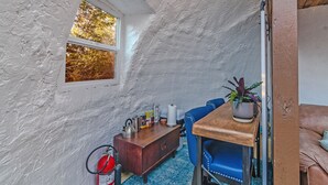Ett litet vindsrum med ett sluttande tak, ett fönster med utsikt över träd, ett träbord med en växt, en blå stol och ett litet skåp med diverse föremål.
