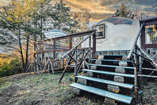 A rustic cabin with a wooden staircase leading to a deck, surrounded by trees.