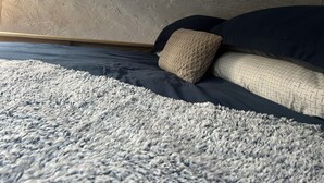 A bed with a dark blue mattress, a beige throw pillow, and a grey shaggy rug.