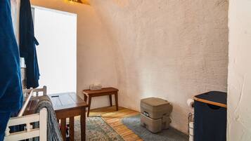 Een kleine, gezellige kamer met houten vloer, een tafeltje, een bankje, een raam met gordijnen en een toilet aan de muur.