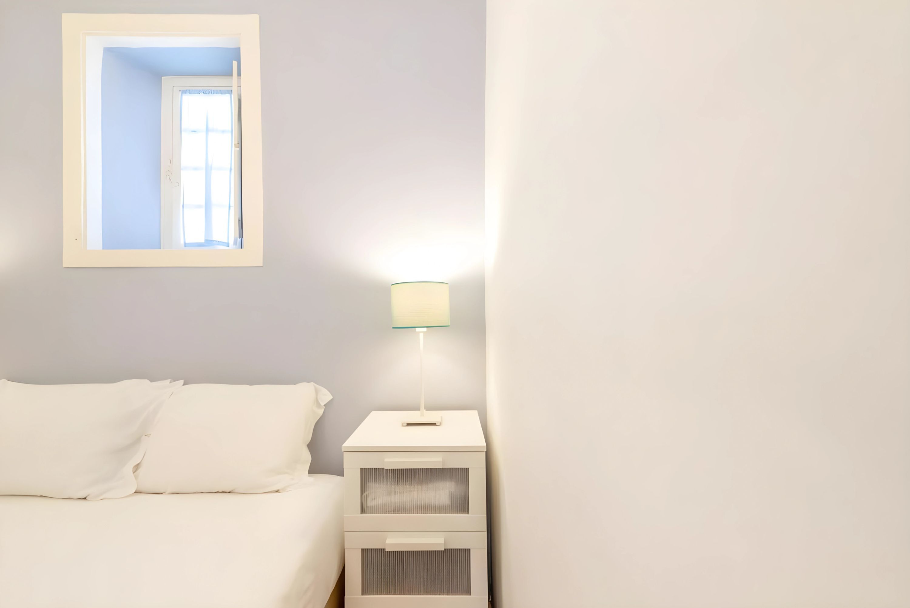 Ein Schlafzimmer mit einem Bett, einem Nachttisch mit Lampe, einem Spiegel und einem kleinen Fenster.