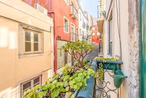 Ein schmaler Stadtgang mit Gebäuden an beiden Seiten, mit Balkonen, die mit Pflanzen und einem grünen Gießkanne geschmückt sind.