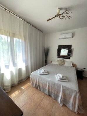 A bedroom with a bed, curtains, an air conditioning unit, a bedside table, and a wall-mounted mirror. - Spacious vacation home for 2–6 people, right on the beach, surrounded by nature, pet-friendly (Sitia)