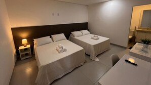 A hotel room with two beds, a desk, a chair, a nightstand with a lamp, and a bathroom visible through an open door. - Flamboyant Stay Rooms (Goiânia)