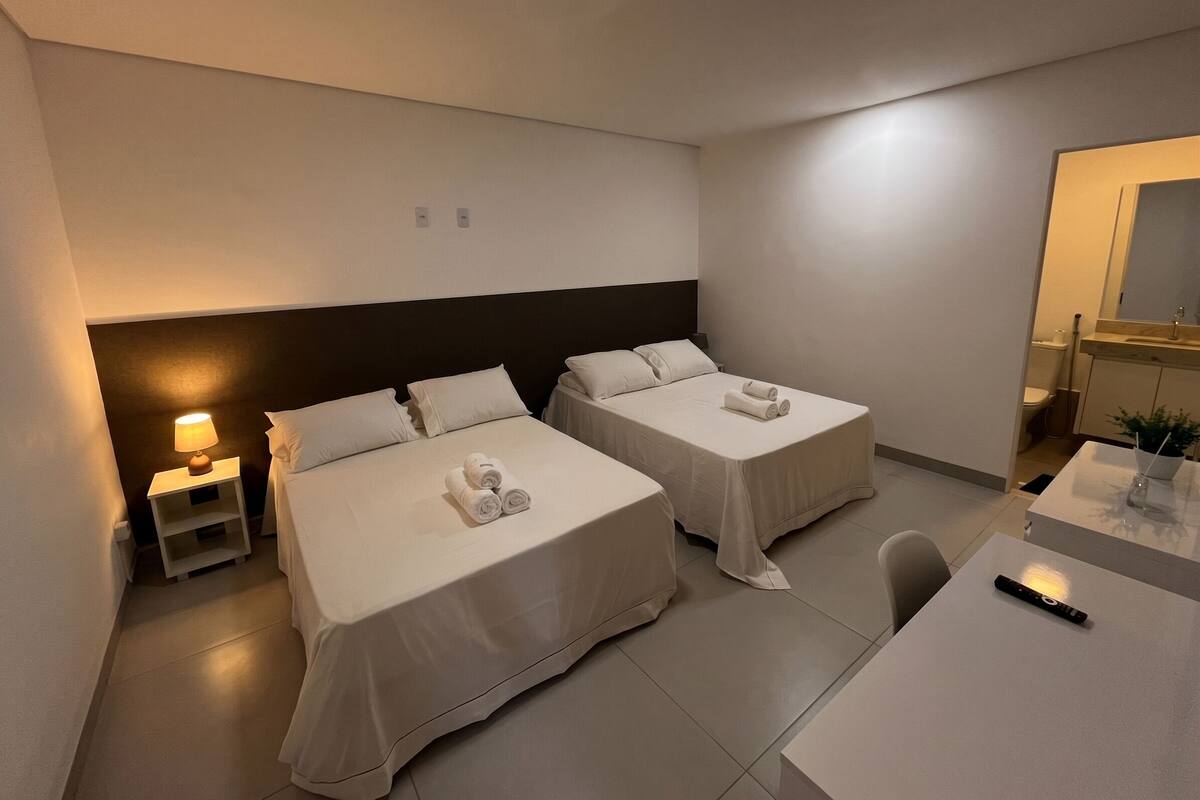 A hotel room with two beds, a desk, a chair, a nightstand with a lamp, and a bathroom visible through an open door.