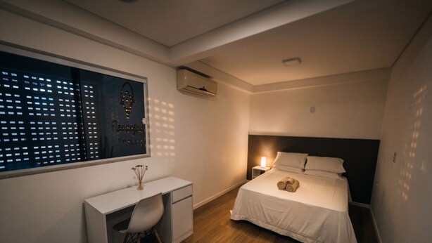 A bedroom with a bed, bedside table, desk, chair, and a window with blinds. - Flamboyant Stay Rooms (Goiânia)