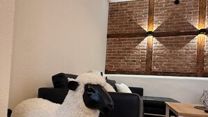 Ein Zimmer mit einer schwarzen Couch, einem weißen, flauschigen Schaf, einem gewebten Teppich und einer Ziegelwand mit zwei Wandleuchten.