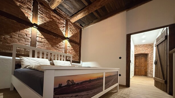 Ein Schlafzimmer mit einem Bett, einem weißen Nachttisch mit Landschaftsbild und einer Holzbalkendecke, sowie einer Wand mit freiliegenden Ziegelsteinen.