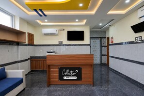 A reception area with a wooden reception desk, a blue sofa, a flat-screen TV mounted on the wall, an air conditioning unit, and a fire extinguisher. - Classic Airport Stay (Nelamangala)