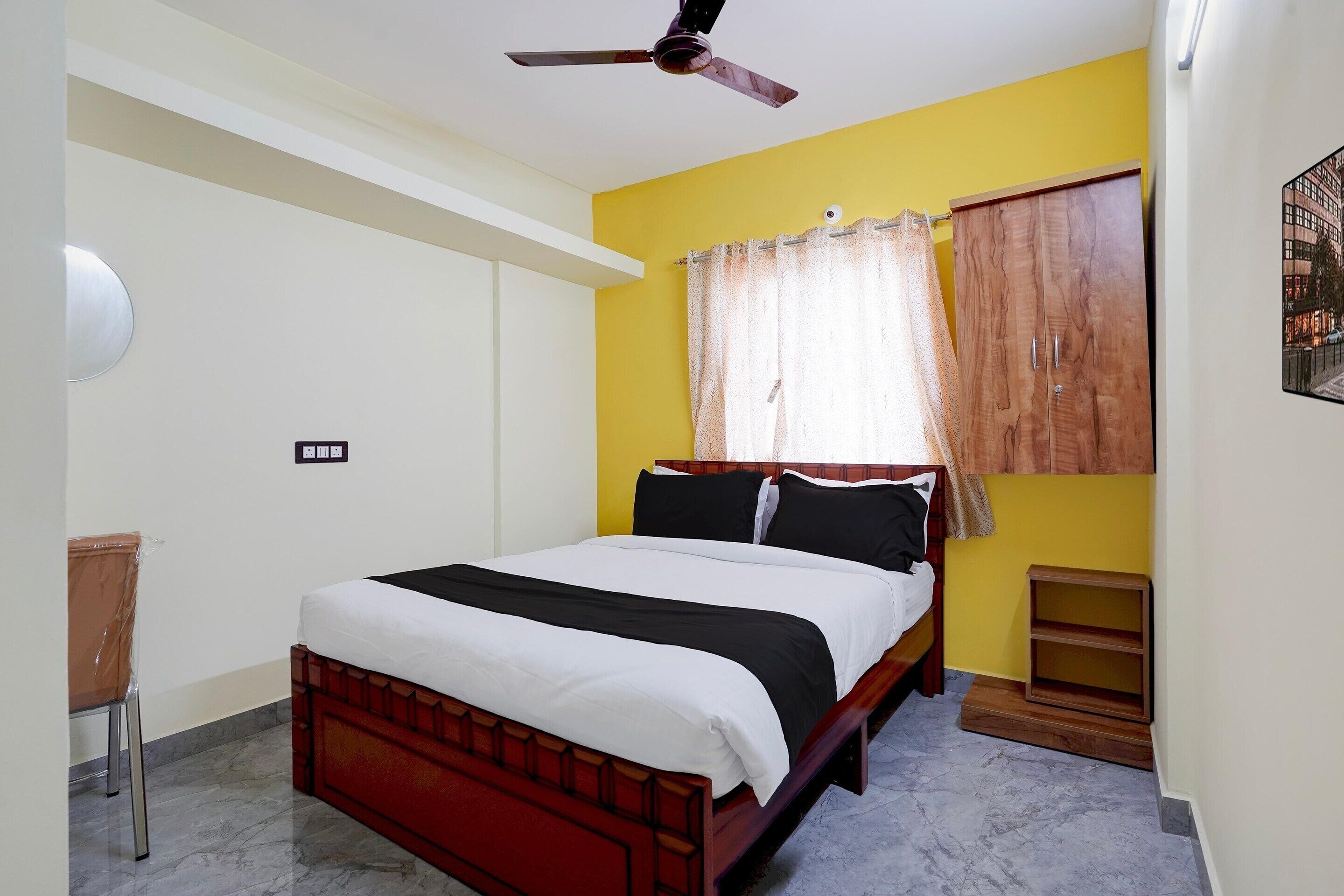 A bedroom with a bed, a ceiling fan, a window with curtains, a wooden wardrobe, and a small bedside table.