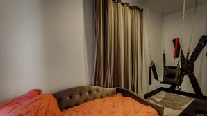 Une chambre à coucher avec une tête de lit capitonnée, une couvre-lit orange, un fauteuil suspendu et un rideau.
