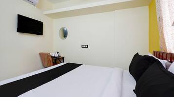 A hotel room with a bed, a TV mounted on the wall, a small table with a chair, and a power outlet on the wall.