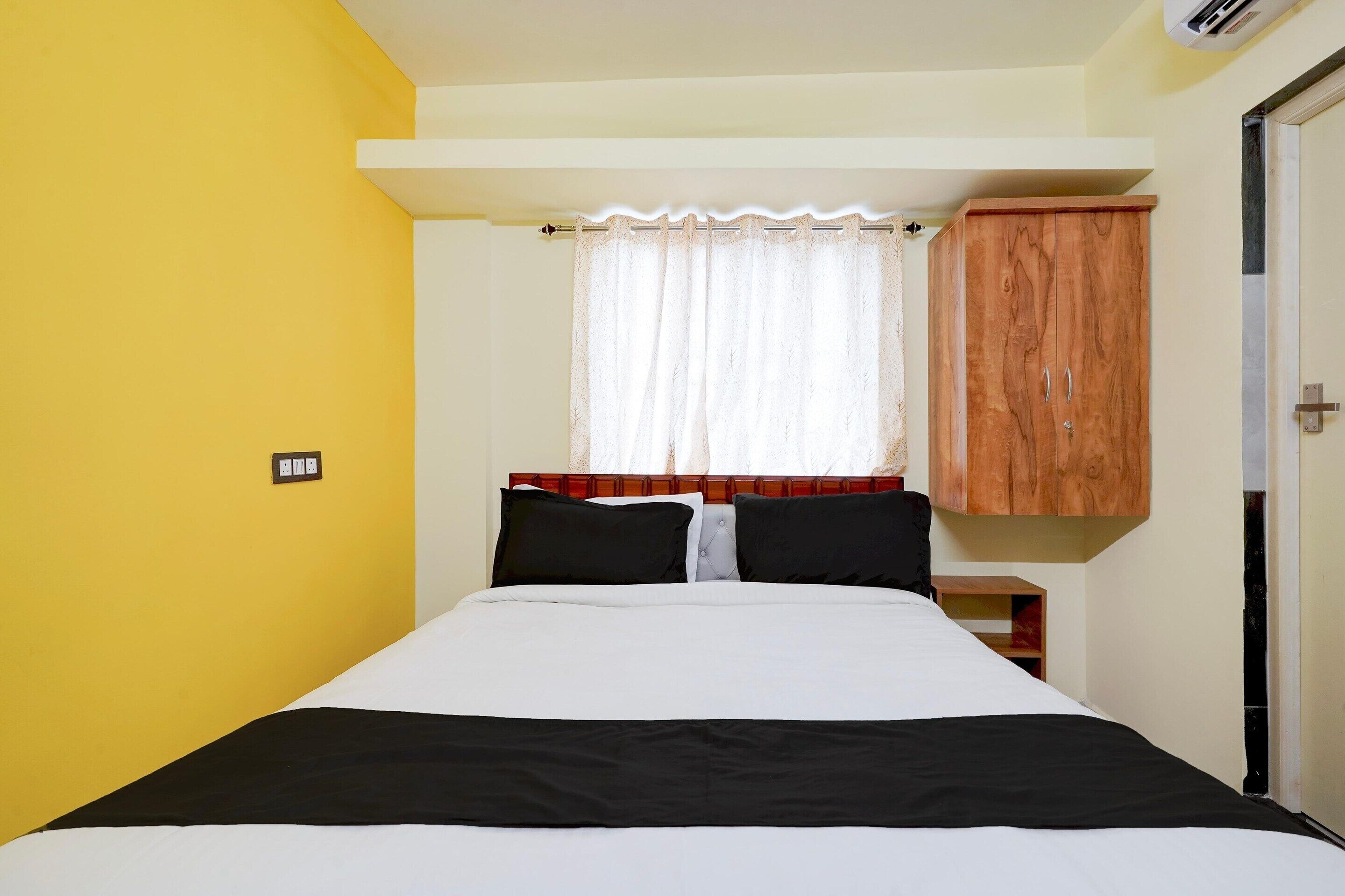 A bedroom with a bed, a wooden wardrobe, a window with a white curtain, and a wall socket.