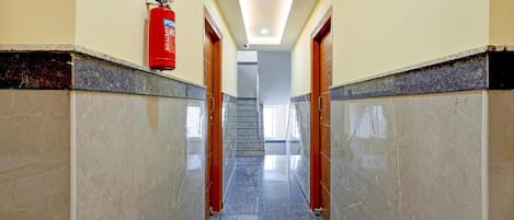 A hallway with a fire extinguisher mounted on the wall, a staircase in the background, and a tiled floor.