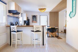 Una cocina moderna con armarios blancos, un extractor de aire negro y una lámpara colgante. Hay dos taburetes blancos y una mesa de comedor con sillas.