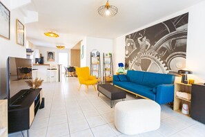 Una sala moderna con un sofá azul, un sillón amarillo y un reposapiés blanco. En el fondo, se observa un comedor con una mesa y sillas.
