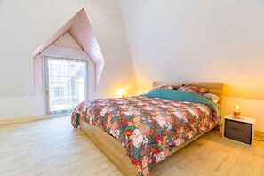 Un dormitorio con techo inclinado, una cama con ropa de cama floral, una mesita de noche con lámpara y una ventana con persianas.