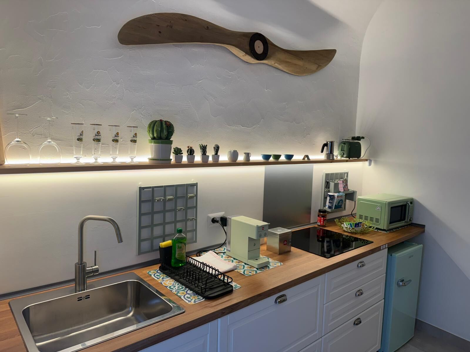 A kitchen with a modern design, featuring a stainless steel sink, a microwave, a coffee maker, and a small refrigerator.