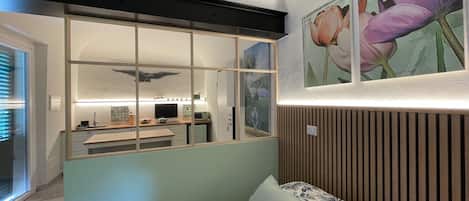 A bedroom with a bed, a window with blinds, a wall-mounted artwork, and a kitchen area visible through a glass partition.